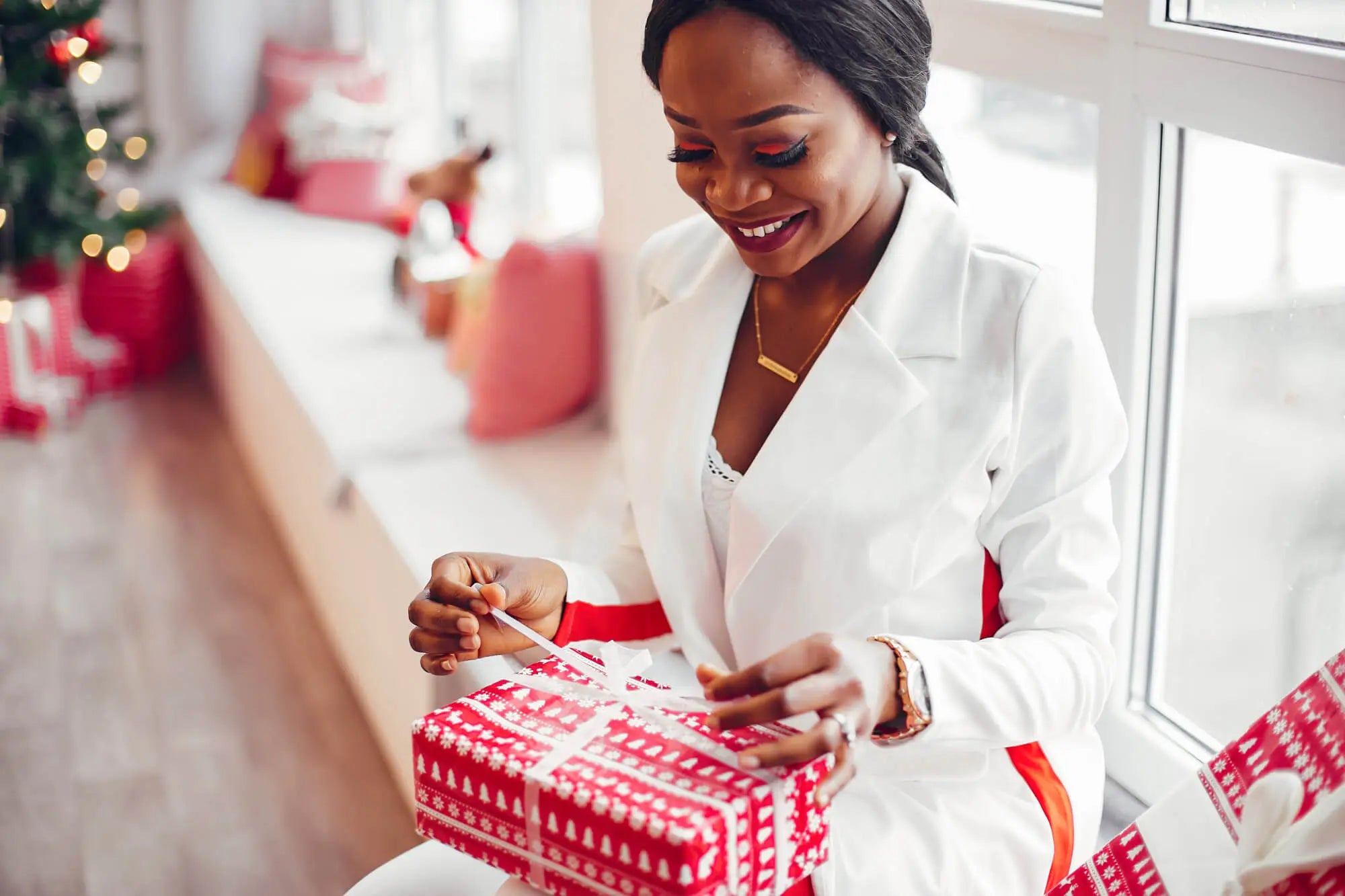 femme noire habillée en blanc souriante, assise au bord de la fenêtre entrain d'ouvrir son cadeau de noël qui contient un parfum