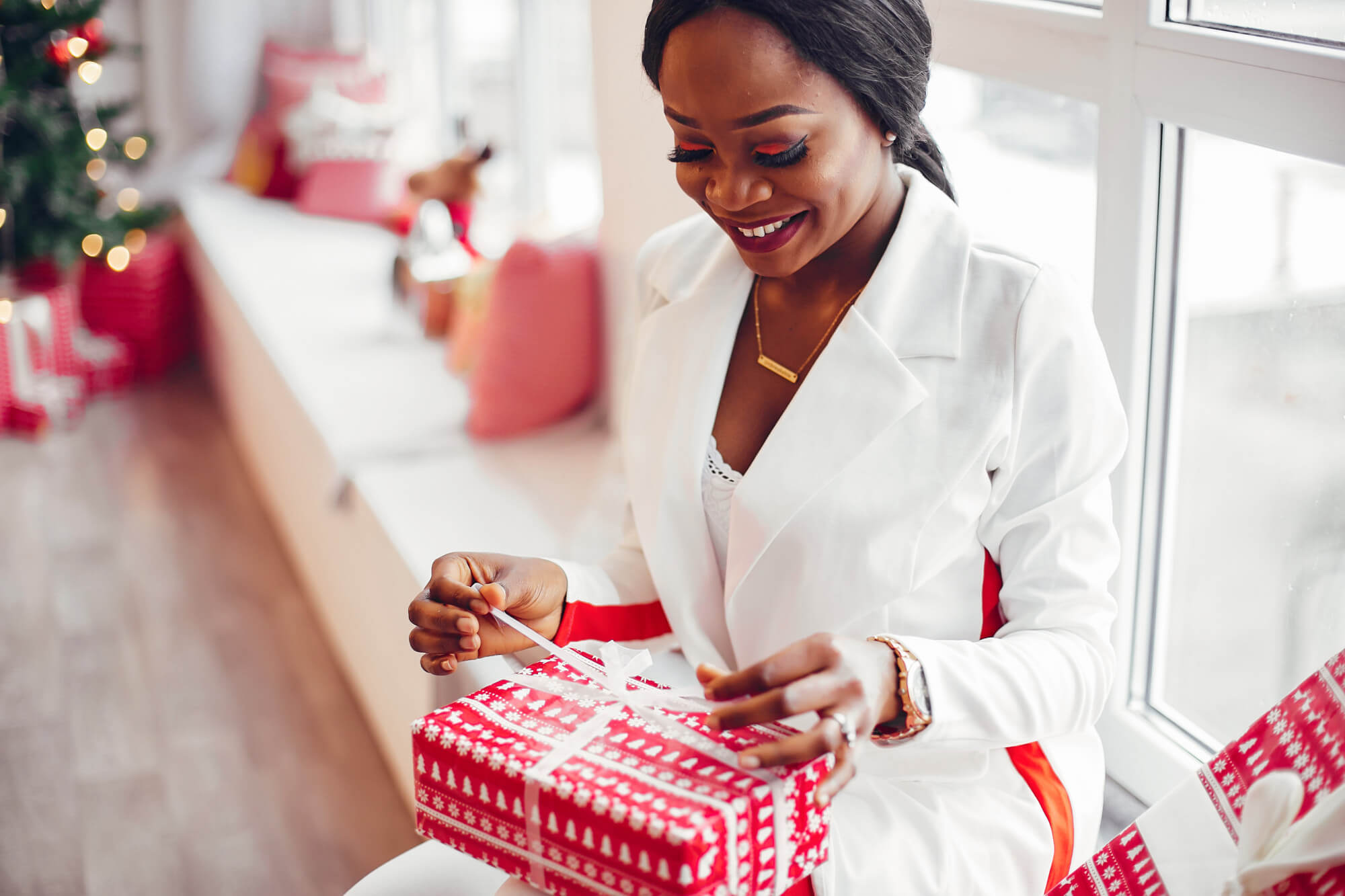 femme noire habillée en blanc souriante, assise au bord de la fenêtre entrain d'ouvrir son cadeau de noël qui contient un parfum